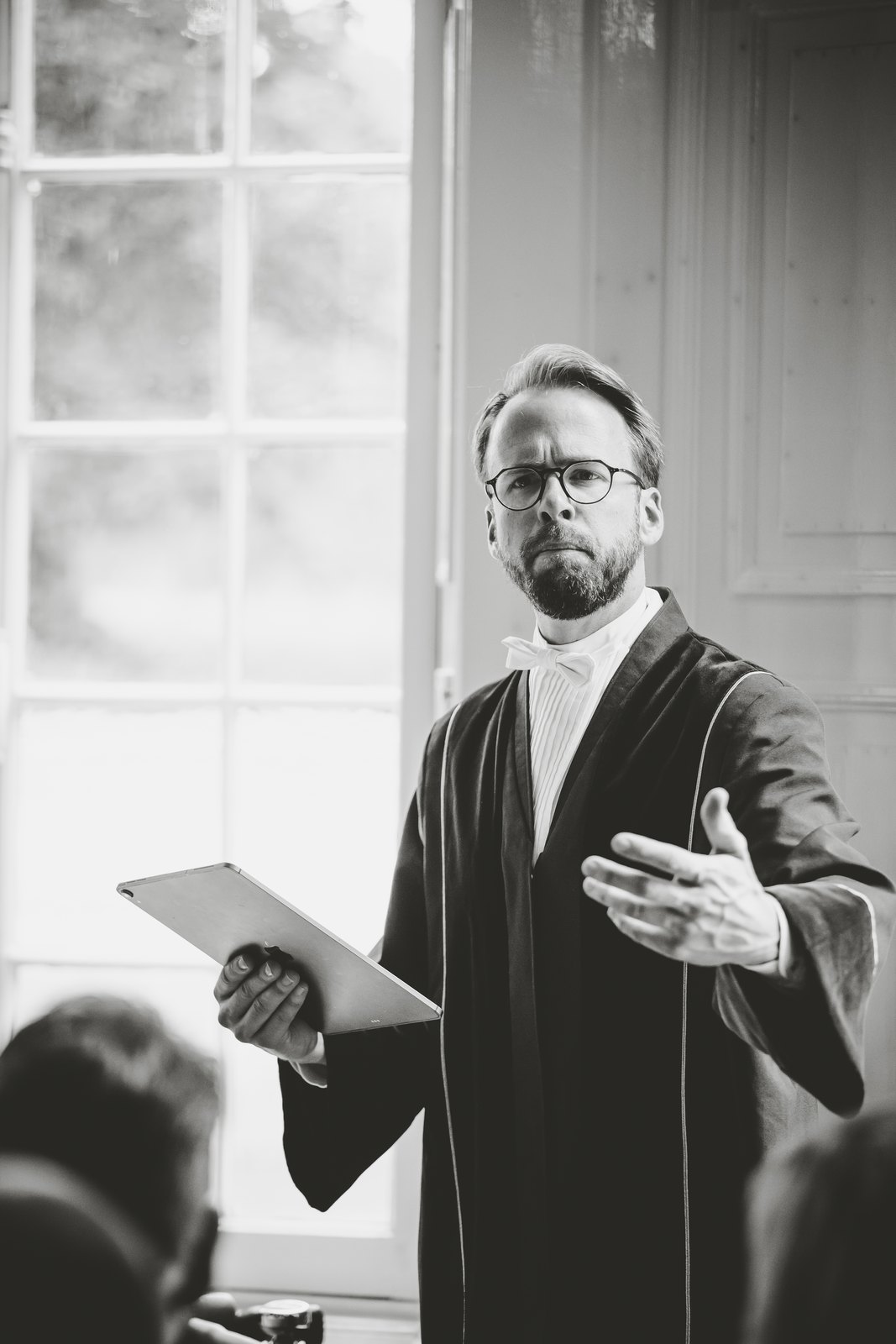Trouwambtenaar Bert Groothedde in toga tijdens een huwelijksceremonie, gericht op de gasten in de zaal